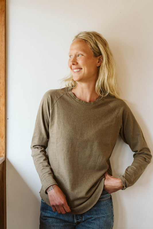 Smiling woman in an olive long sleeve tee and jeans stands near a sunny window