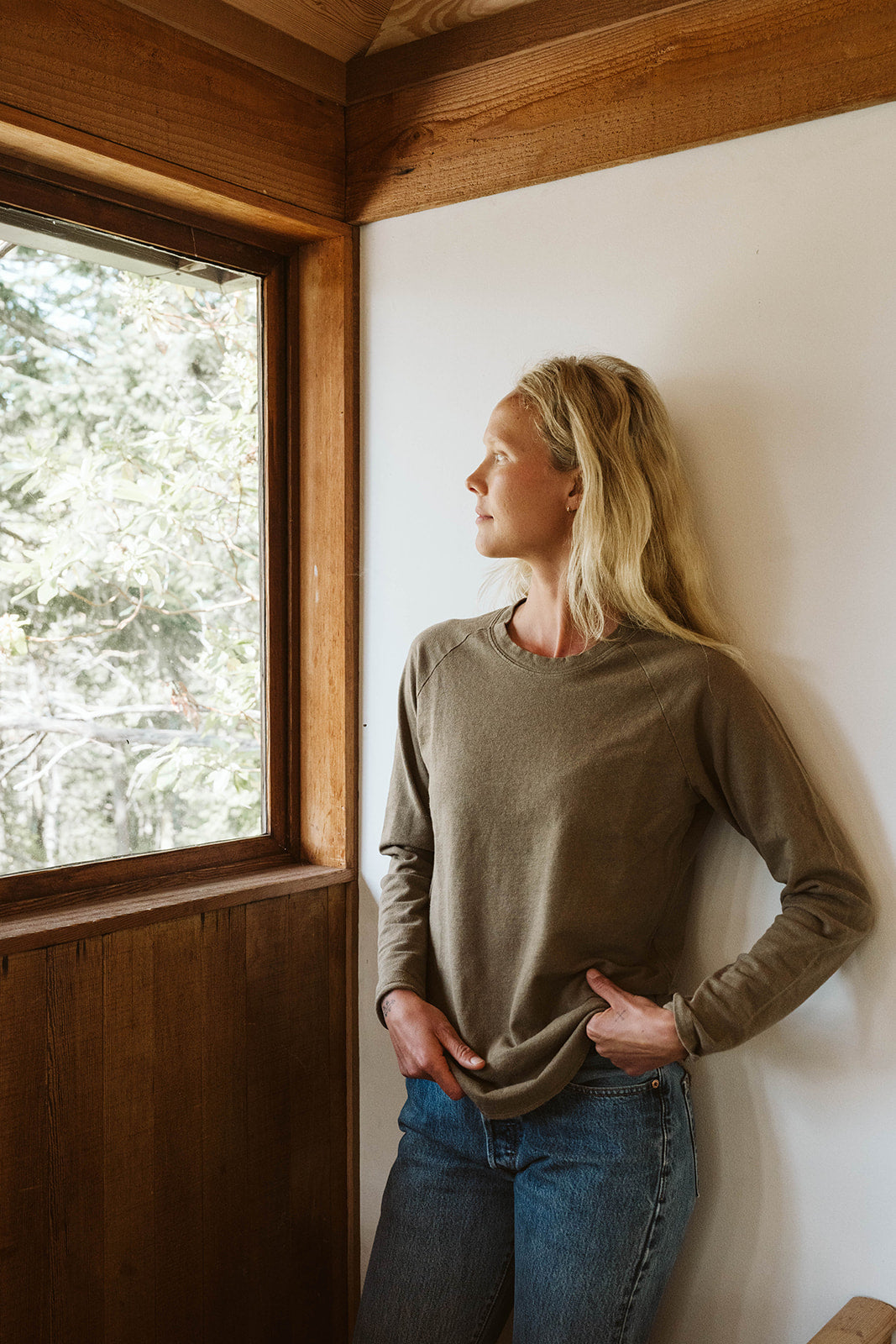 Woman in olive long sleeve shirt gazes thoughtfully out a window in a wood-accented room